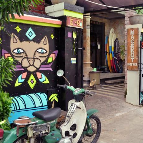 Entrance of Macskot Hostel in Bali with a painted cat mural, surfboards and a green scooter parked outside.