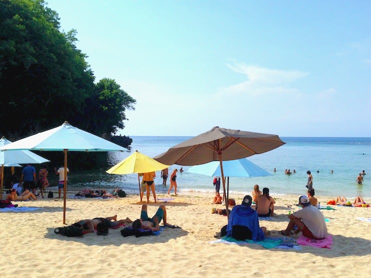 Sunbathers under umbrellas on the sand at Padang Padang Beach, Bali, with swimmers in the sea.