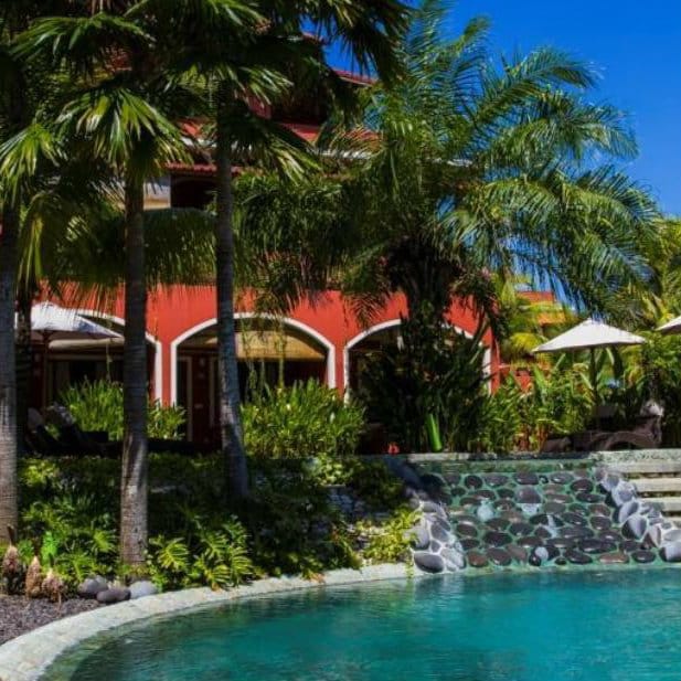 Red villa building framed by palm trees beside a turquoise pool at Pinkcoco in Bali.