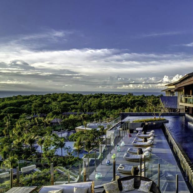 Rooftop pool deck with loungers overlooking forest and the sea at RIMBA in Jimbaran, Bali.