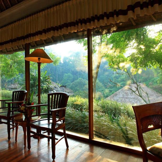 Bali villa lounge with wooden chairs and a lamp beside a full-height window over misty jungle.