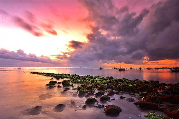 Pink and orange sunrise over mossy rocks and fishing boats at Sanur beach, Bali.