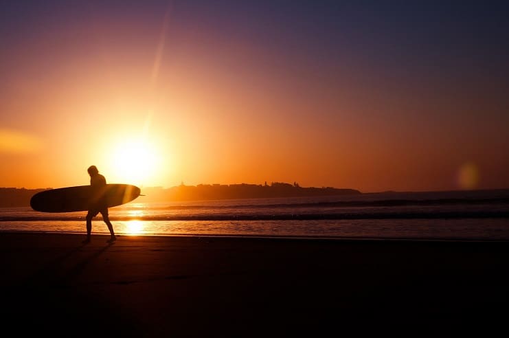 Surfer silhouetted carrying a longboard along the beach at sunset in Canggu, Bali.