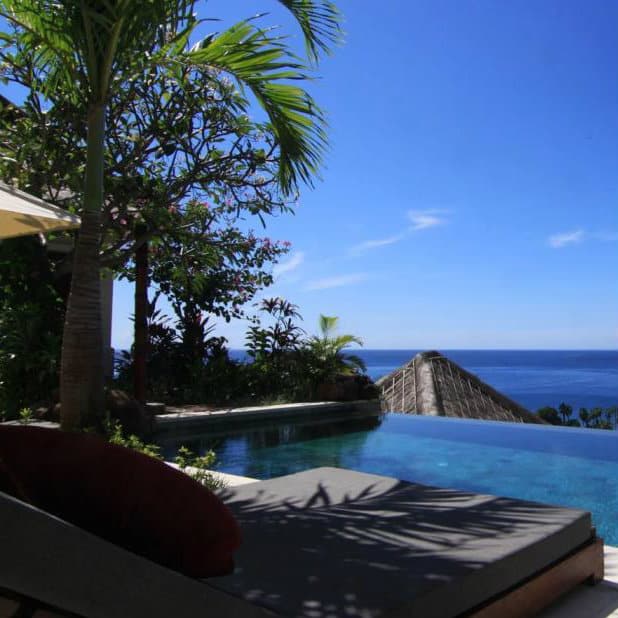Clifftop infinity pool and daybed with a red cushion overlooking the ocean at a Bali villa.