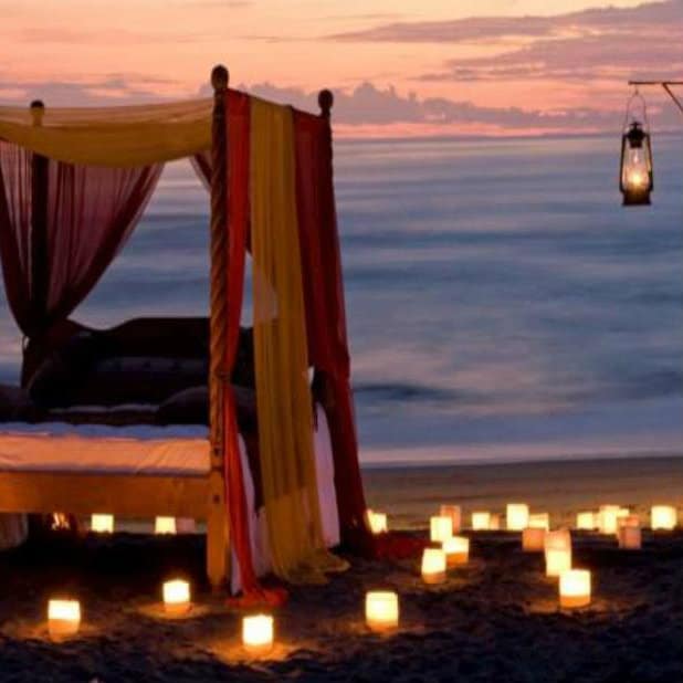 Draped four-poster bed on the beach lit by candles and a lantern at sunset in Bali.