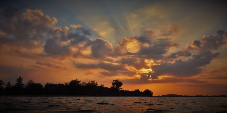 Sun rays breaking through clouds at sunset over a calm sea with a wooded shoreline in Indonesia.