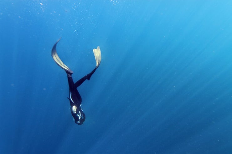Freediver in a wetsuit descending head-first through deep blue water in Bali, light rays above.