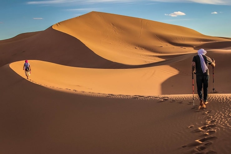 Two hikers cross golden Sahara sand dunes in Morocco, one wrapped in a blue headscarf.