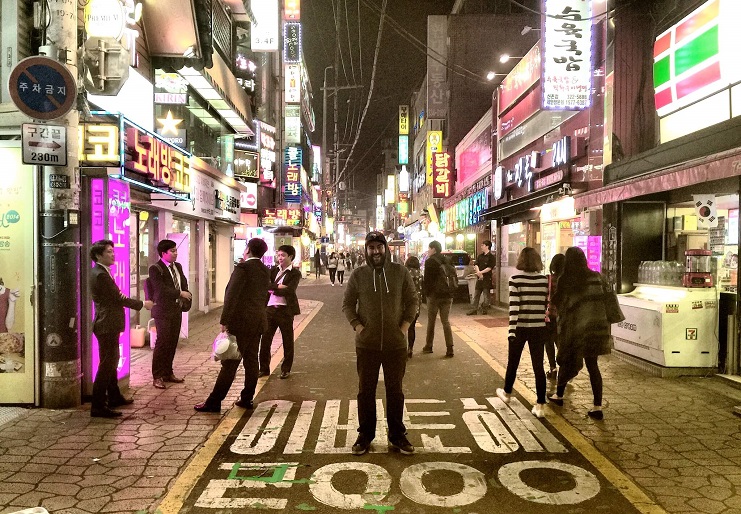 Alice stands on a neon-lit shopping street in South Korea at night, crowds passing by.