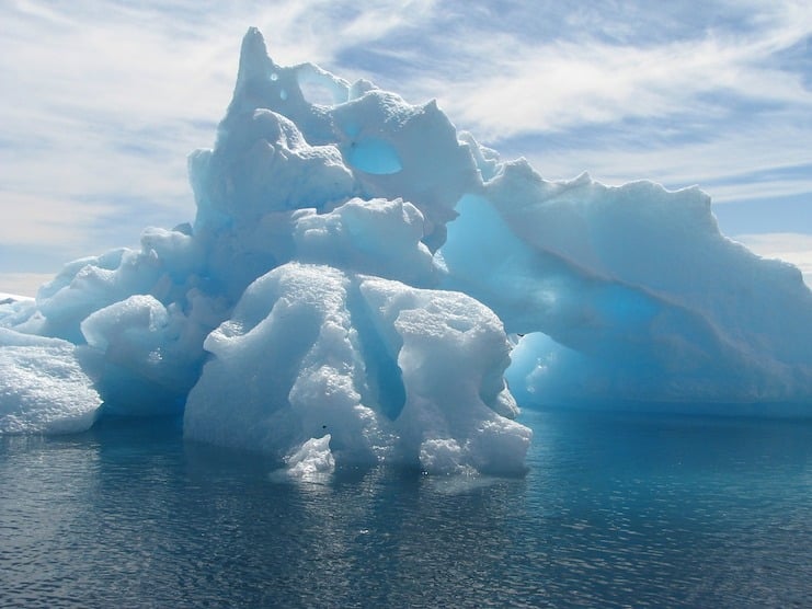 A jagged blue and white iceberg floating on calm Antarctic water under a hazy sky.