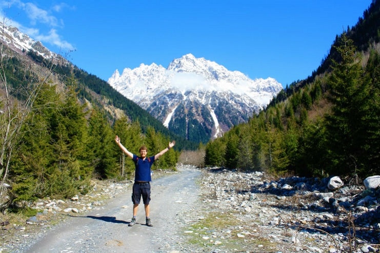 A hiker raises both arms on a mountain trail with snowy peaks and pine forest ahead.
