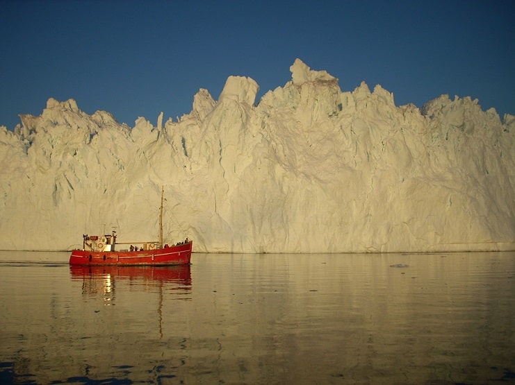 A small red fishing boat drifts past a towering white iceberg near Ilulissat, Greenland.