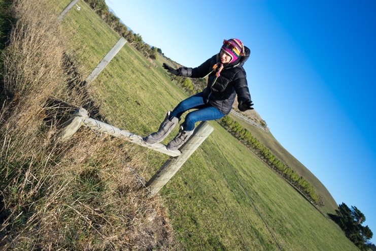 Alice sits on a wooden fence in a grassy field wearing a helmet, photo tilted sideways.