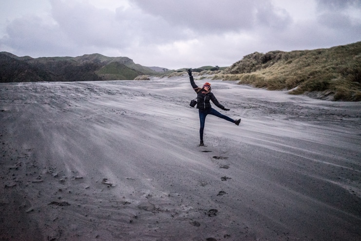 Alice balances on one leg on a windswept grey sand beach under a cloudy sky.