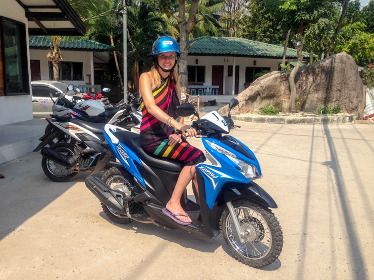 Alice grins in a helmet while sitting on a blue rental scooter outside guesthouse rooms.