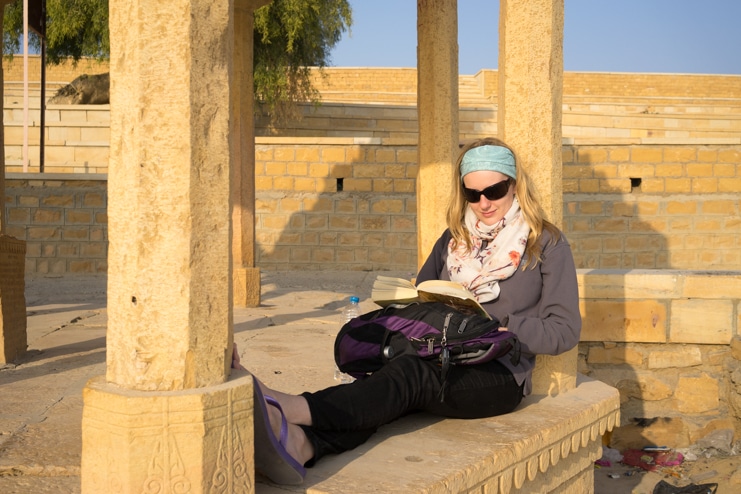 Alice reads a book while sitting in the shade of a sandstone pavilion in sunglasses.