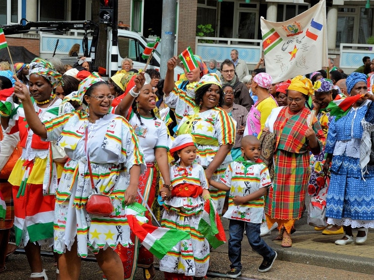 Women and children in bright striped Surinamese dress wave flags at a Keti Koti street parade.