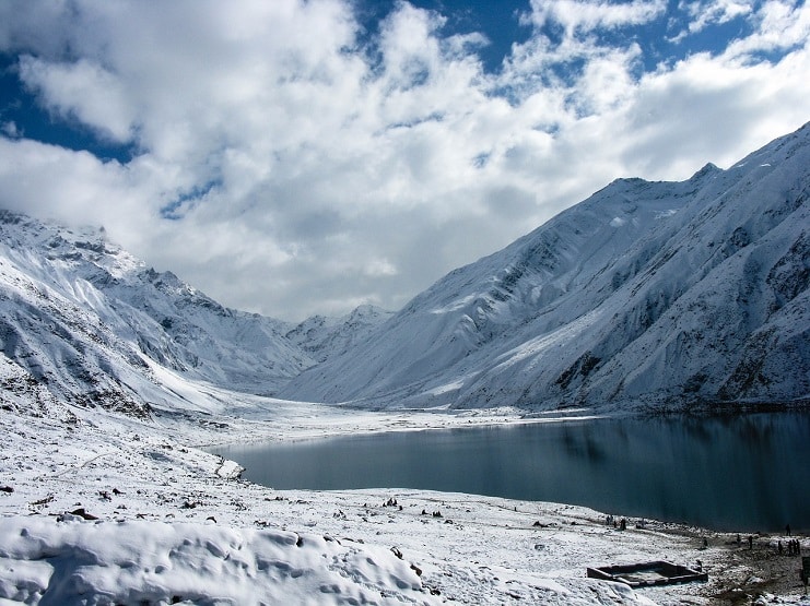 A dark alpine lake between snow-covered mountains under a cloudy sky.