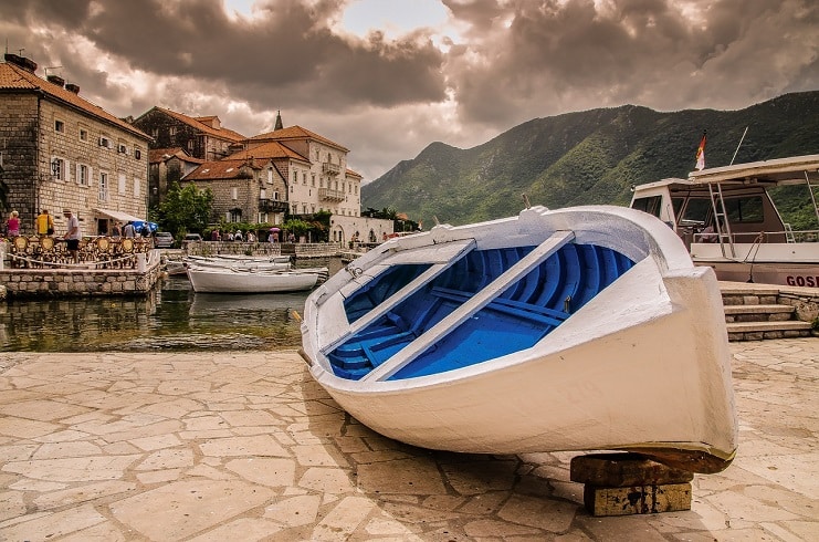 A white and blue rowing boat sits on the stone quay of Perast, Montenegro, with mountains behind.