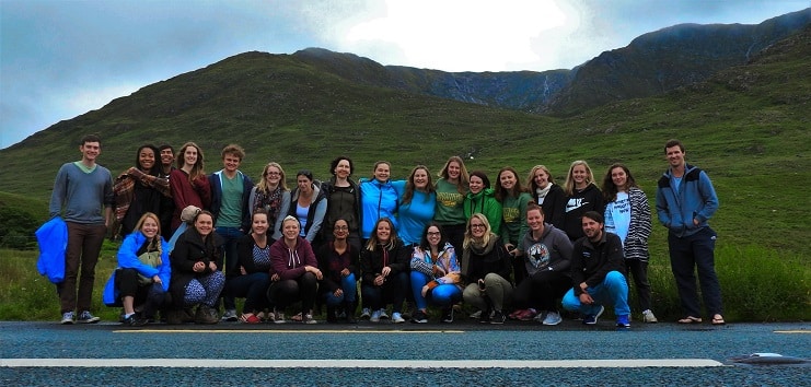 A large group of young travellers pose on a road below green Connemara mountains in Ireland.
