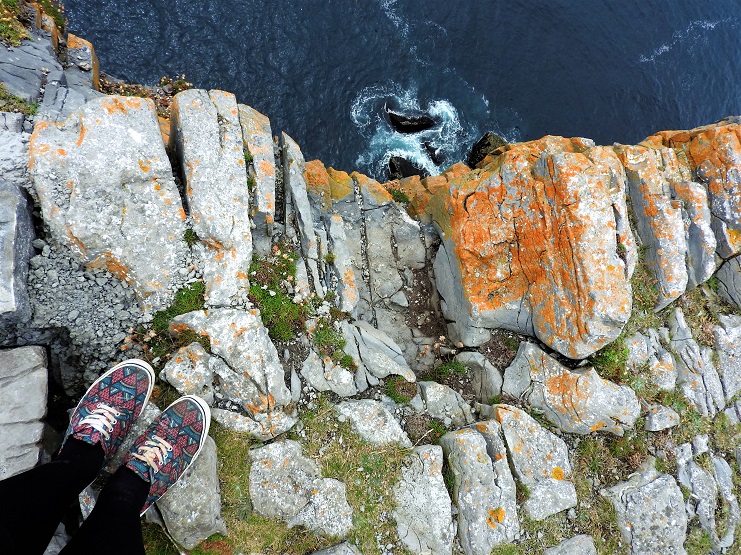 Looking down from a lichen-covered clifftop in Ireland to waves breaking on rocks far below, Alice's feet at the edge.