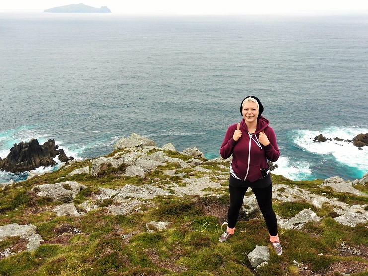 Alice gives two thumbs up on a grassy clifftop above the sea near Dingle, an island offshore.