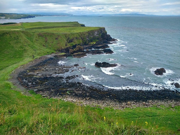 Green cliffs and a rocky bay along the Antrim coastline in Northern Ireland under grey sky.