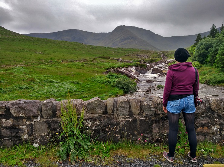 Alice stands on a stone bridge looking out over a stream and misty green mountains in Connemara.