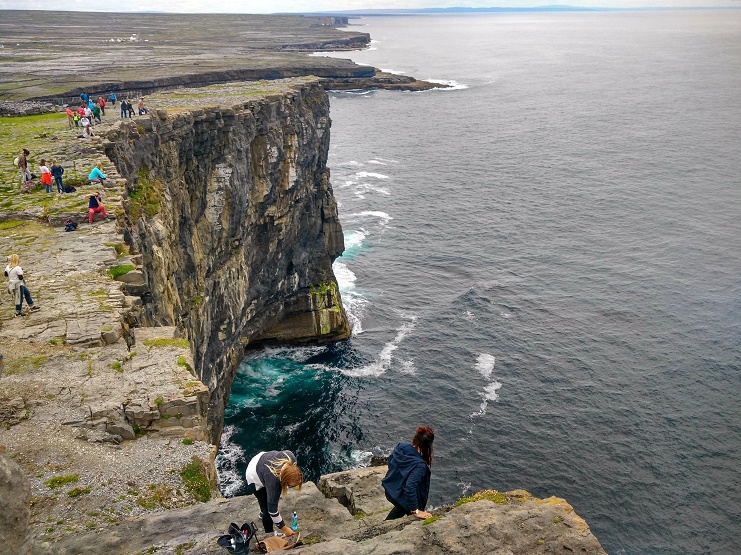 People sit at the edge of the sheer cliffs at Dun Aengus on the Aran Islands, waves below.