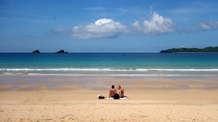 A couple sits on the sand looking out over a bright blue sea in El Nido, Palawan.