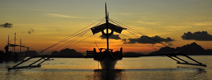 An outrigger boat silhouetted on calm water at sunset in El Nido, Palawan.