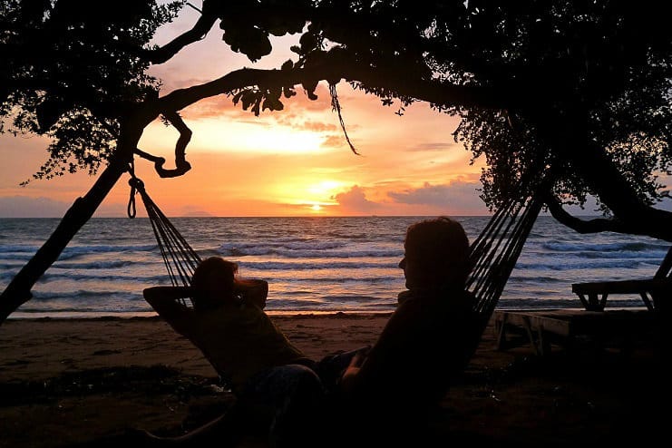 Two people relax in hammocks under a tree watching the sunset over the sea.