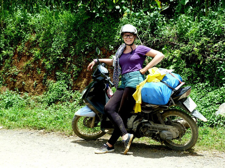 Alice in a helmet leans against a loaded motorbike on a roadside beside dense green foliage.