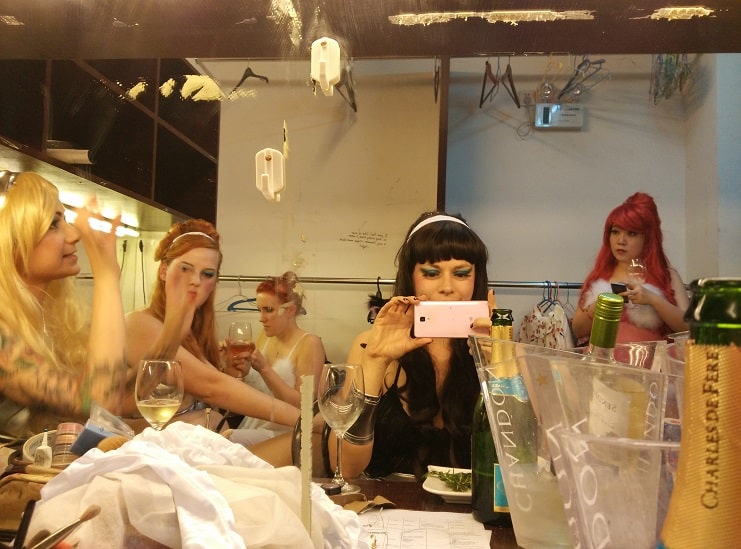 Burlesque performers getting ready in a mirrored dressing room with wine and champagne.