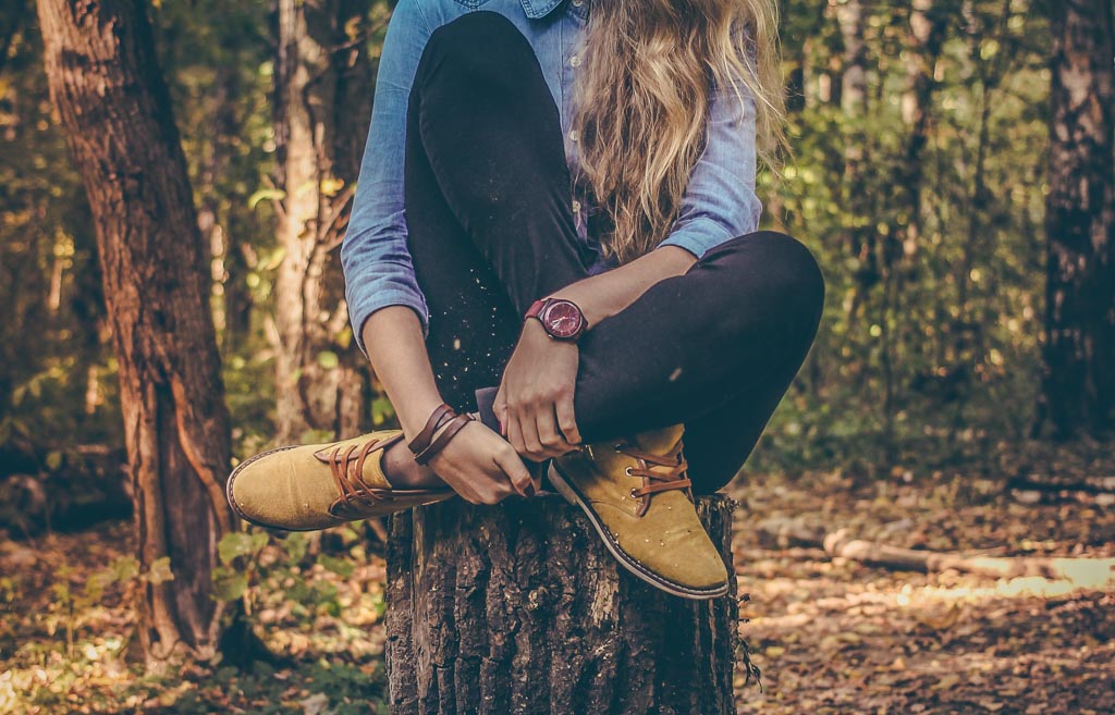 Woman in denim shirt and mustard boots sitting cross-legged on a tree stump in autumn woodland.