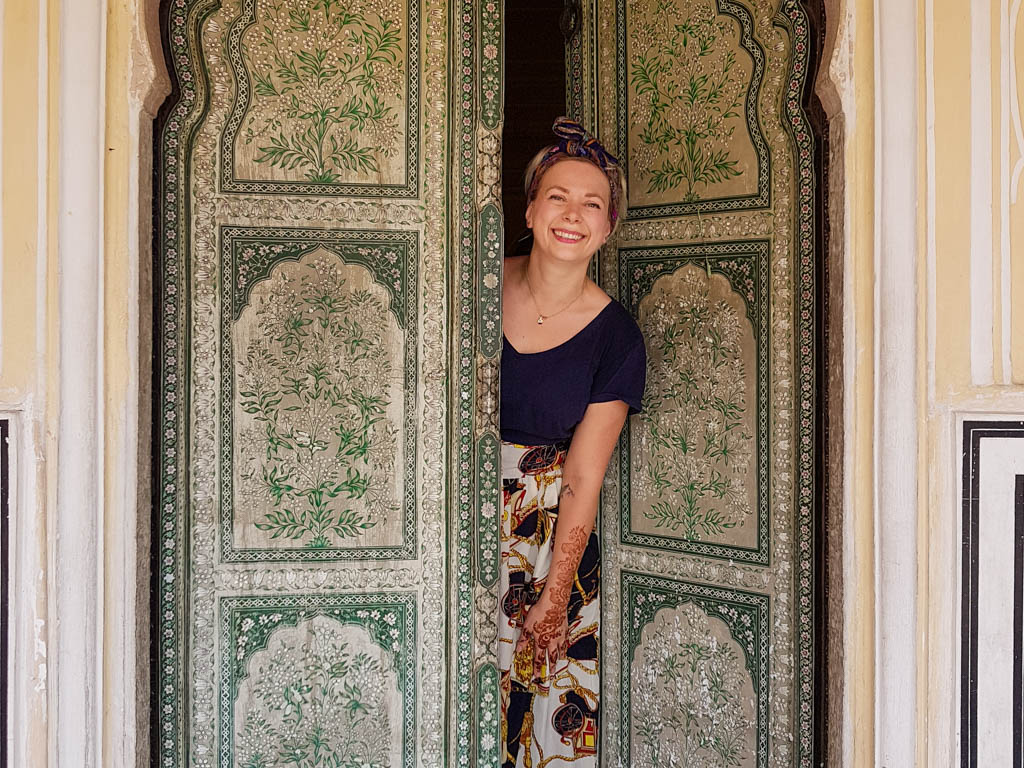 Alice smiles in a painted palace doorway in Jaipur, India, wearing a navy top and printed skirt.