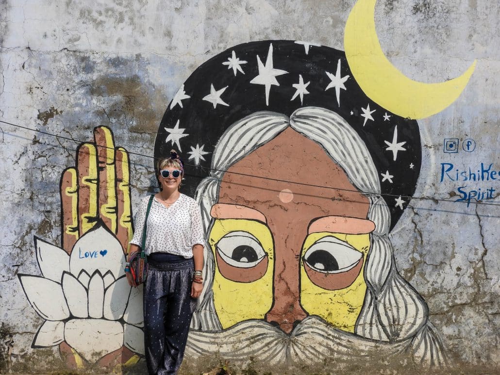 Alice standing beside a Rishikesh wall mural of a bearded face, crescent moon and lotus in India.