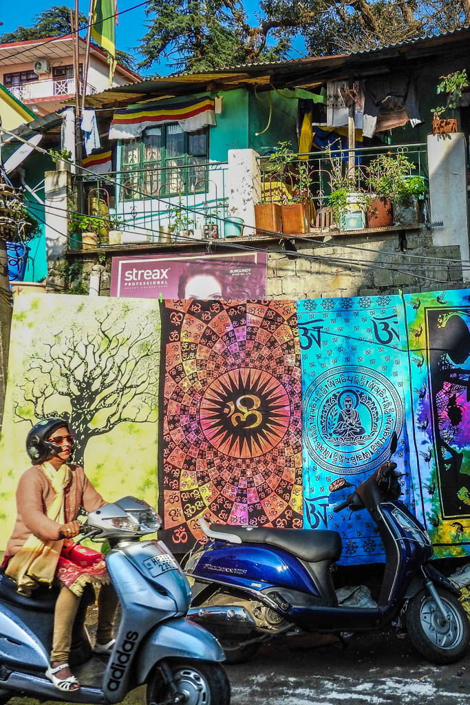 A woman rides a scooter past patterned tapestries hung for sale on an Indian street.