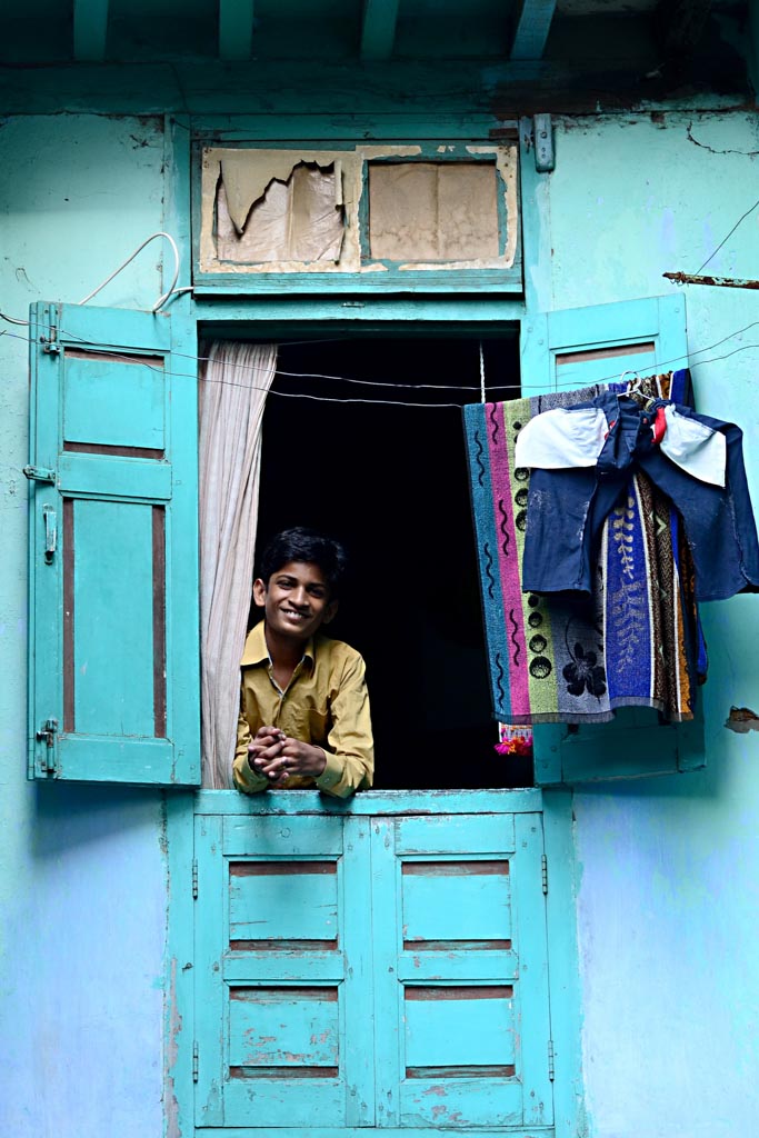 A smiling young man leans out of a turquoise painted window in India, laundry hung beside it.