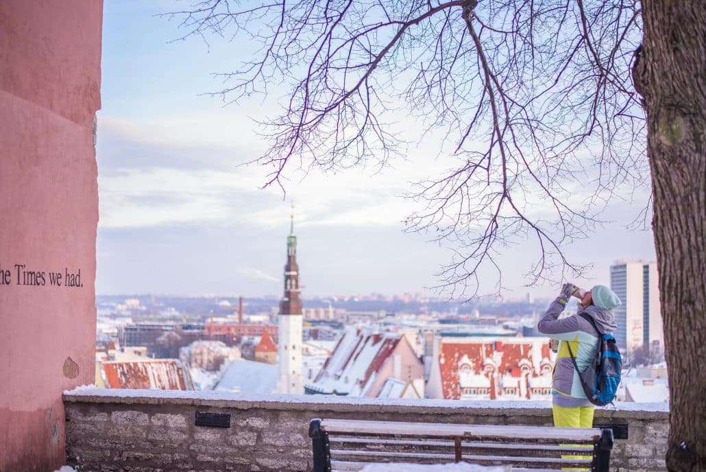 A person drinks from a bottle overlooking snowy rooftops and a church spire in wintry Tallinn, Estonia.