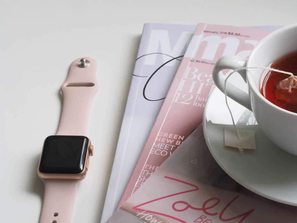 A pink smartwatch and stacked magazines beside a cup of tea with the bag steeping.