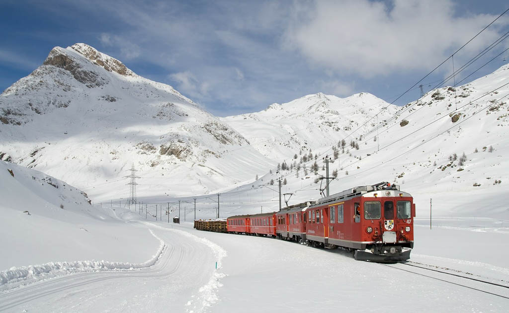 A red train winding through a snow-covered valley beneath rocky mountains under a blue sky.