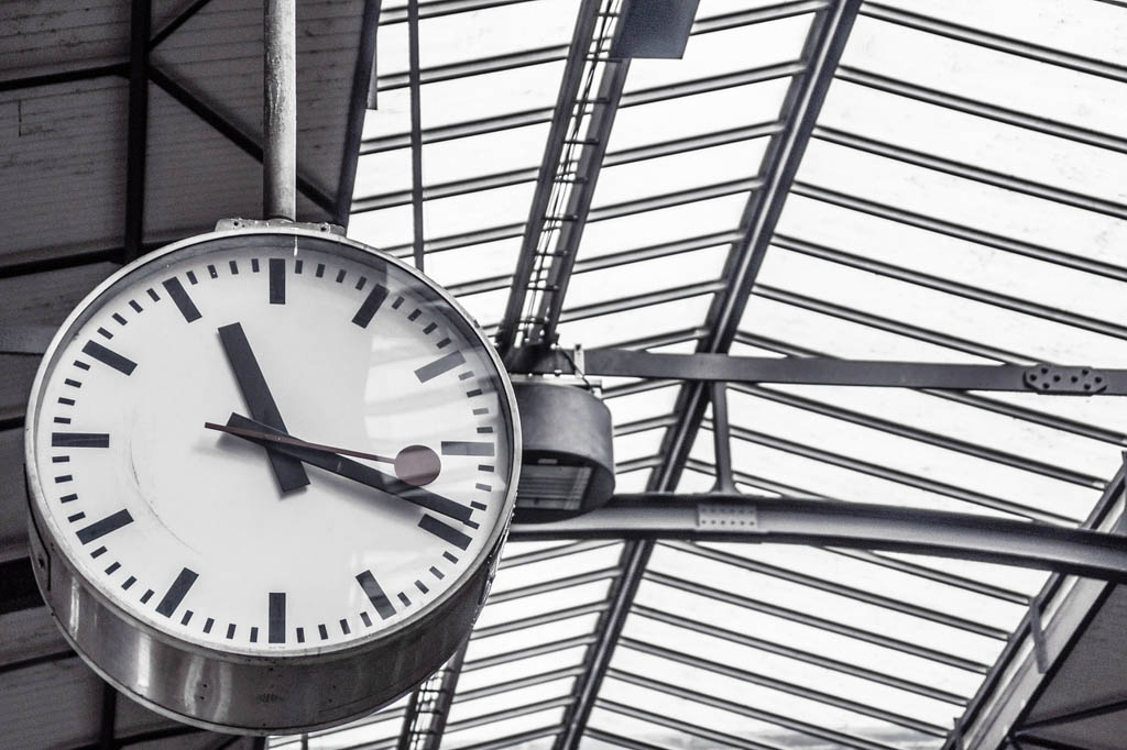 A large white station clock hanging beneath the glass and steel roof of a train station.
