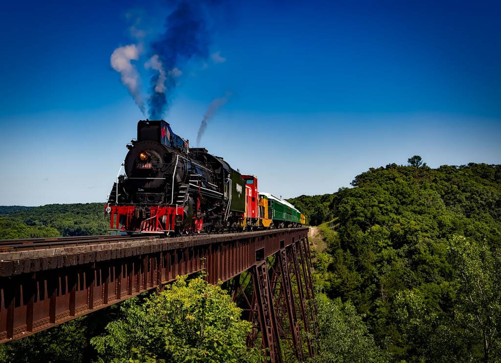 A black steam locomotive with colourful carriages crossing a high metal trestle bridge over forested hills.
