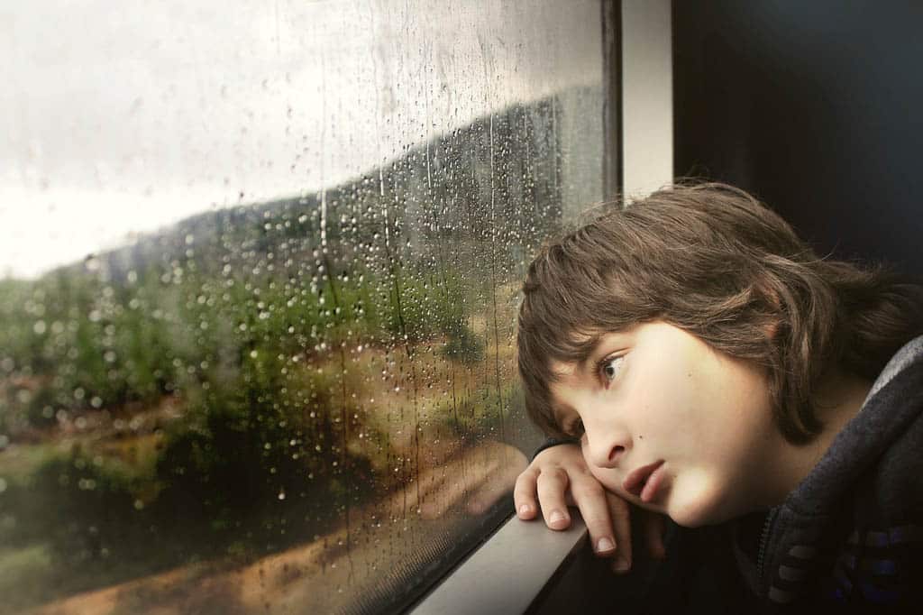 A boy resting his head on his arm, gazing out of a rain-streaked train window at green countryside.