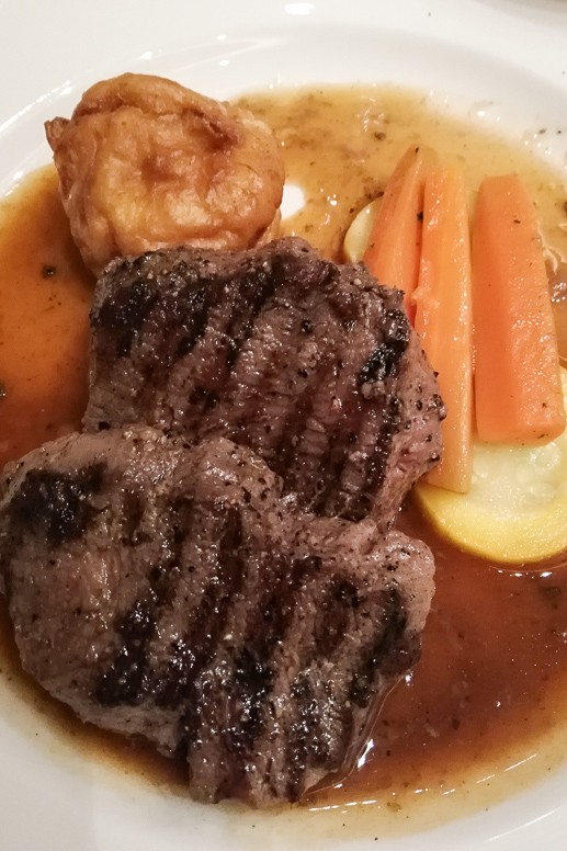 Grilled steak with carrots, potatoes and a Yorkshire pudding in gravy on a white plate.