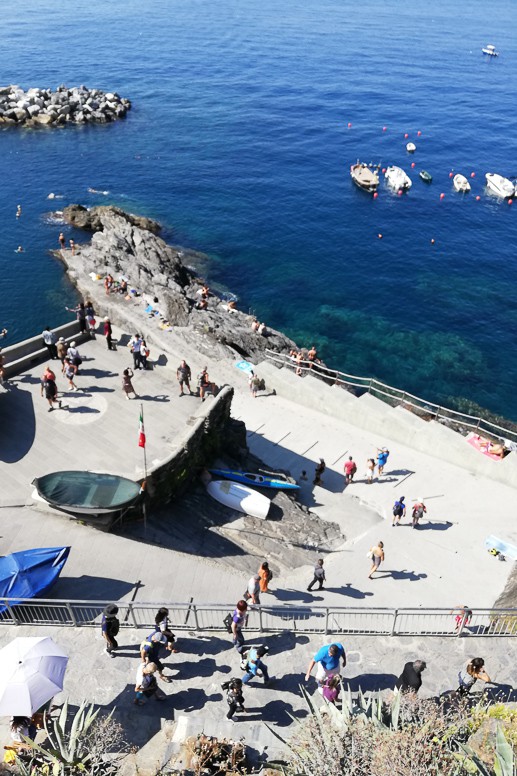 Overhead view of a rocky harbour with moored boats and people walking the waterfront in Cinque Terre, Italy.