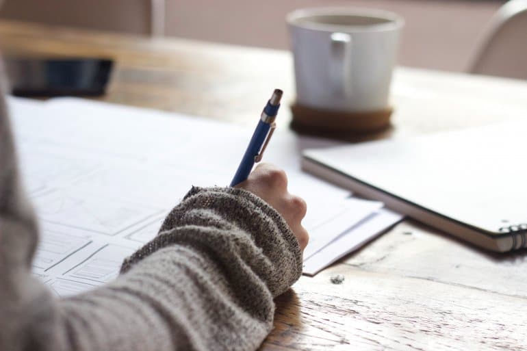A person in a knitted jumper writes with a blue pen on paper at a wooden desk beside a mug of coffee.