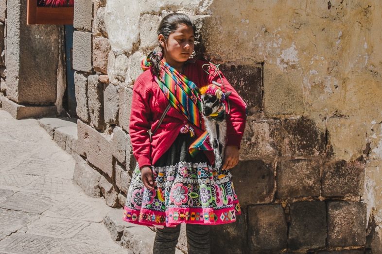 A girl in a red jacket and embroidered skirt holds a lamb against a stone wall in Cusco, Peru.
