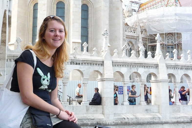Alice sits smiling in front of a white Gothic church facade with tourists behind her.